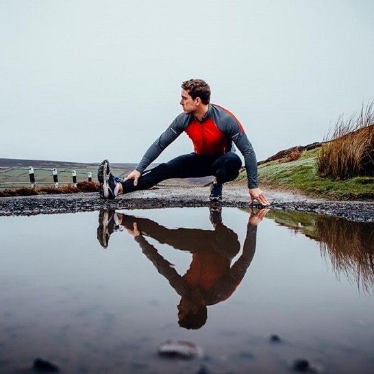 Male runner training outdoors in wet weather. His reflection seen in a puddle of water before him. He stretched his leg.